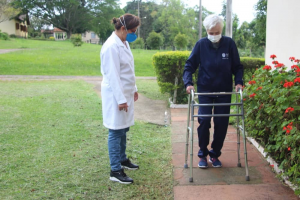 Pessoa idosa anda com auxílio de andador em jardim, enquanto uma profissional da saúde observa